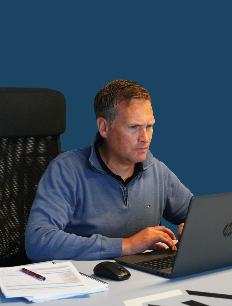 A man in a blue jumper working at a laptop on a desk, with papers and a notebook beside him.
